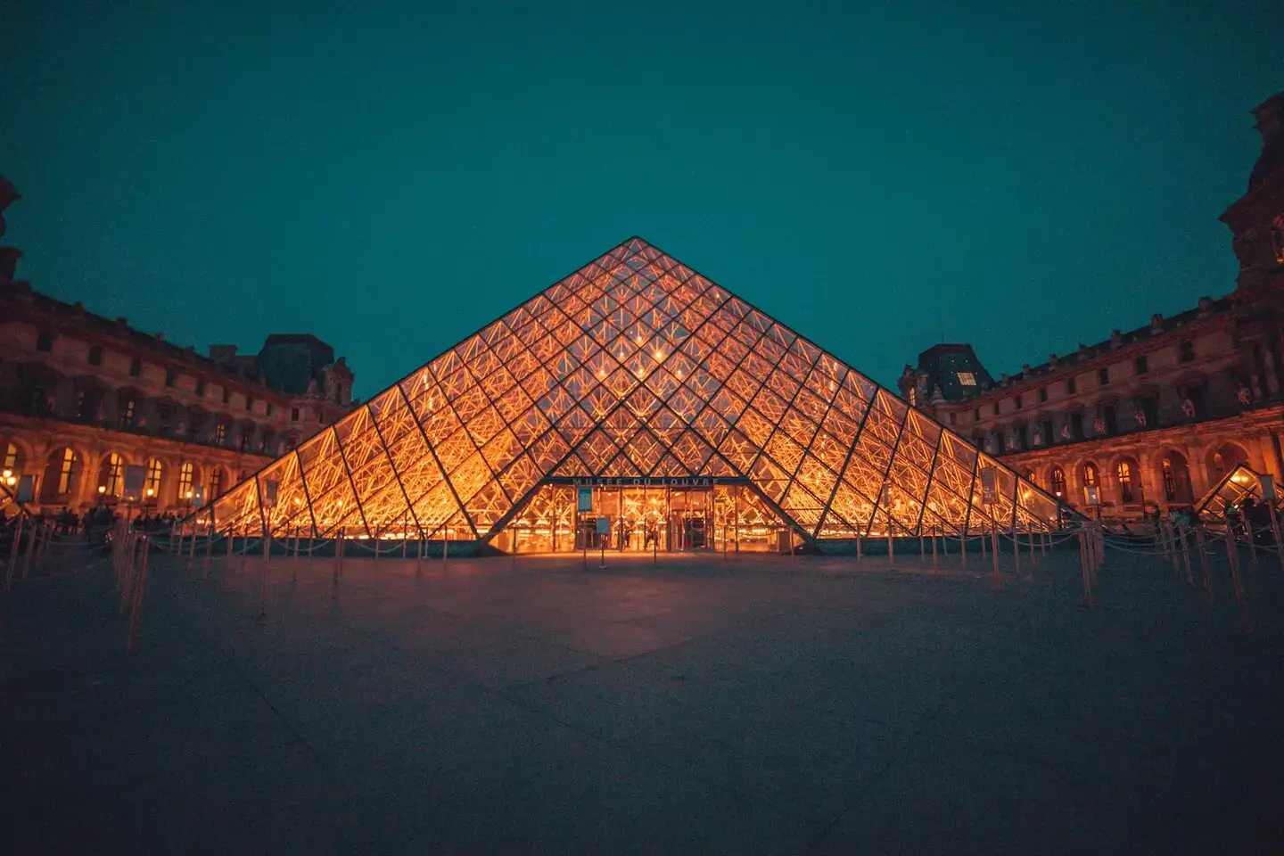 Uma vista noturna da iluminada Pirâmide do Louvre em Paris, cercada pela escuridão e as estruturas históricas do museu