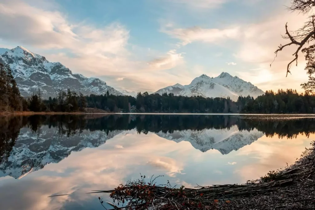 Lago refletindo as montanhas nevadas com arvores em tons de marrom escuro, a água parece intocavel